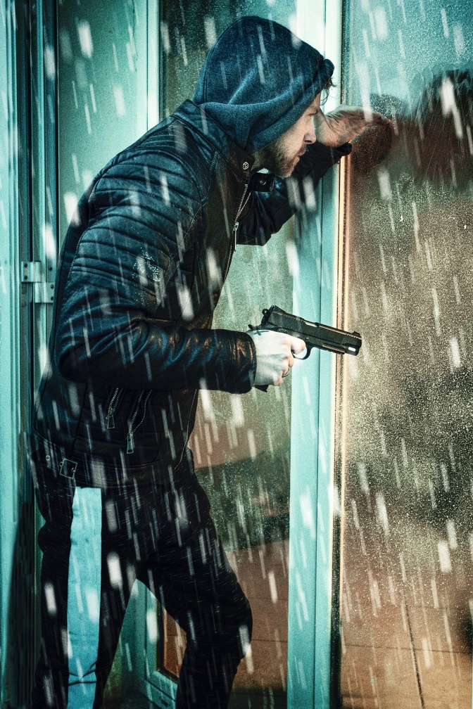 Ein Mann in schwarzer Kleidung steht im Regen mit einer gezückten Waffe vor einer Glastür.