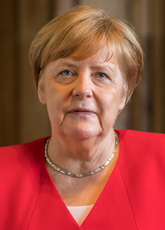 Angela Merkel mit blonder Föhnfrisur und ernstem Blick in einem roten Blazer.