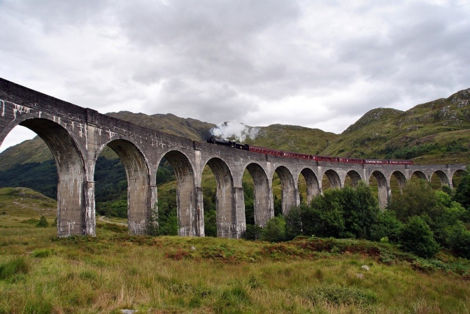 Ein Zug mit Dampflok fährt über eine hohe Brücke durch einen schottische Landschaft