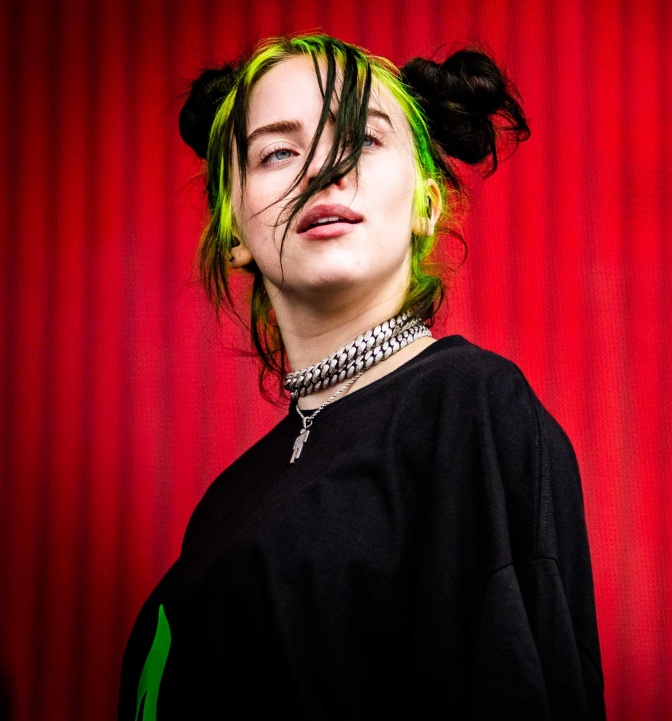 Billie Eilish mit Haaren in schwarz und neongrün, vor einem roten Vorhang.