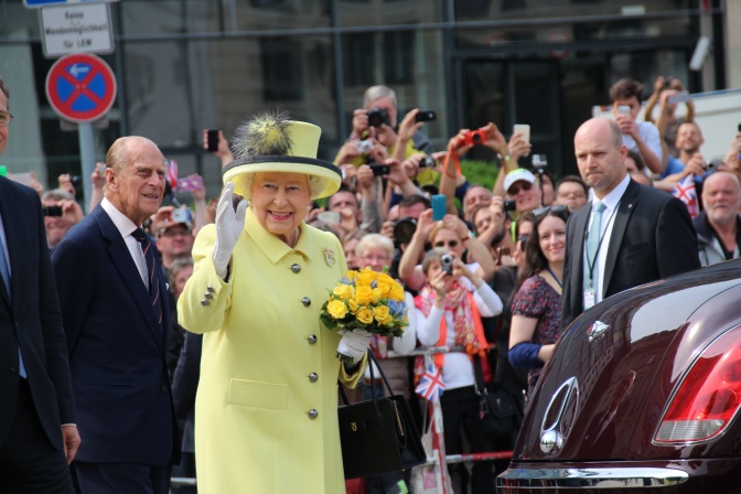 Die Queen winkend in einer Menschengruppe. Sie trägt ein hellgelbes Kostüm mit passendem Hut.