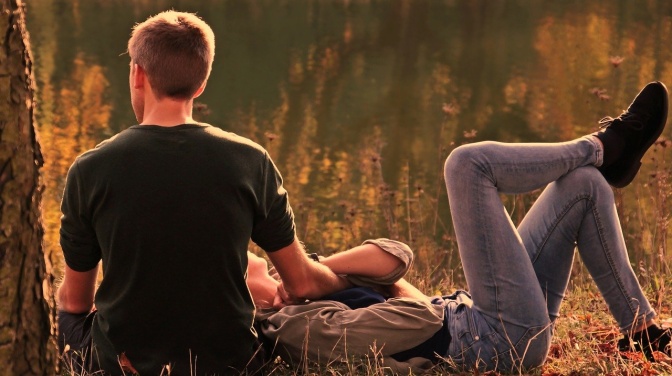 Ein Paar im Freien, er sitzt, sie liegt vor ihm auf einer Wiese, beide halten Händchen.