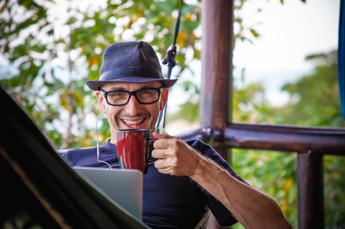 Ein Mann arbeitet am Laptop und hat Kopfhörer in den Ohren. Er lächelt und trinkt eine Tasse Kaffee.