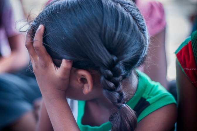Ein Mädchen im Grundschulalter mit geflochtenen Zöpfen versteckt ihr Gesicht hinter ihrer Hand.