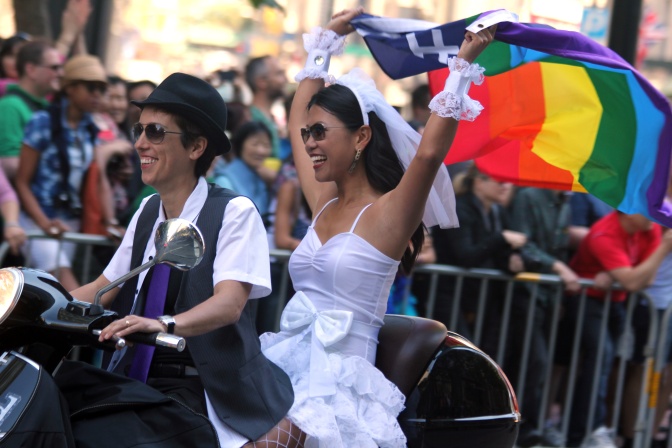 Eine Frau in maskuliner Kleidung fährt ein Motorrad, eine Frau in Brautkleid und Schleier sitzt hinter ihr und hält eine Regenbogenflagge hoch. Am Straßenrand stehen Menschen.