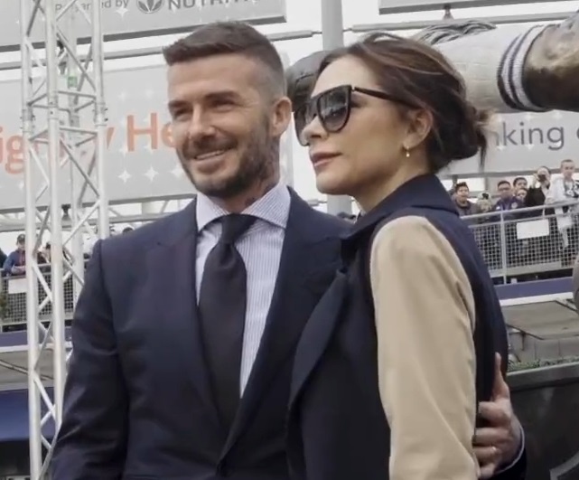 David und Victoria Beckham stehen nebeneinander, er legt den Arm auf ihren Rücken. Er lächelt, sie nicht. Sie trägt eine große, dunkle Sonnenbrille.