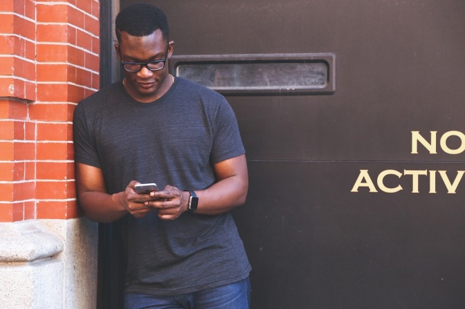 Ein schwarzer Mann steht vor einer Metalltür und nutzt sein Handy.