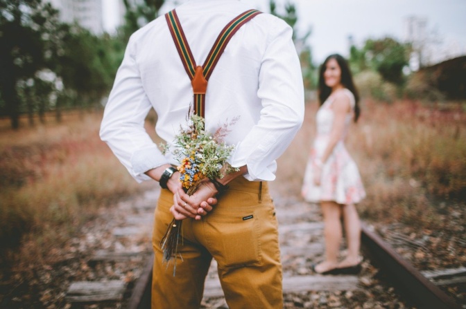 Ein Mann geht auf eine Frau zu und versteckt dabei einen Blumenstrauß hinter seinem Rücken.
