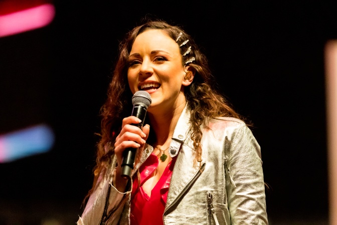 Jasmin Wagne mit langen Locken und vielen Haarspangen im Haar. Sie steht auf der Bühne und singt in ein Mikro.