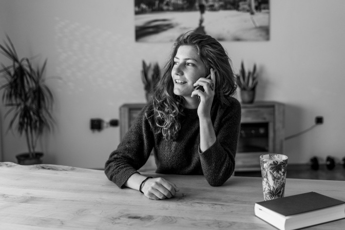 Schwarz-weiß Foto einer Frau, die am Tisch sitzt und mit dem Handy telefoniert. Neben ihr stehen eine Kaffeetasse und ein Buch auf dem Tisch.