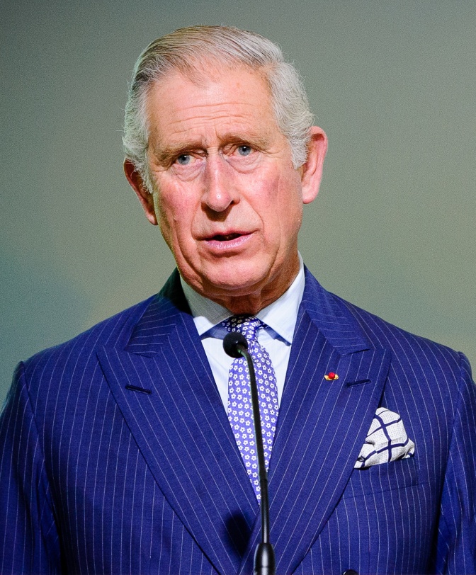 Prinz Charles in einem Nadelstreifenanzug und mit Krawatte und Einstecktuch. Er hat weiße Haare und spricht in ein Mikrophon.