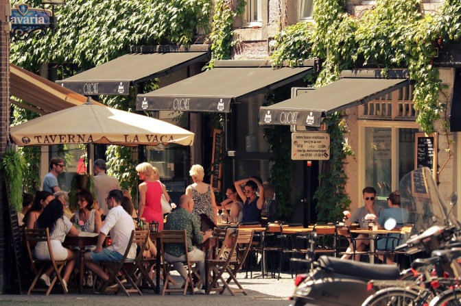 Menschen sitzen auf der Straße vor einem Café, zum Teil unter Sonnenschirmen. Andere Menschen gehen daran vorbei.