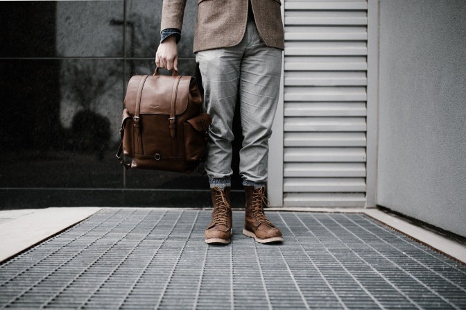 Ein Mann steht vor einem heruntergelassenen Rollladen. In der Hand hält er einen mittelgroßen Lederrucksack.