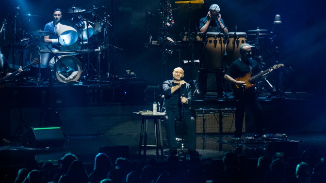 Phil Collins auf der Bühne. Er sitzt vor einem Mikrophon, die Bühne ist bläulich beleuchtet. Hinter ihm sieht man andere Musiker und Instrumente.