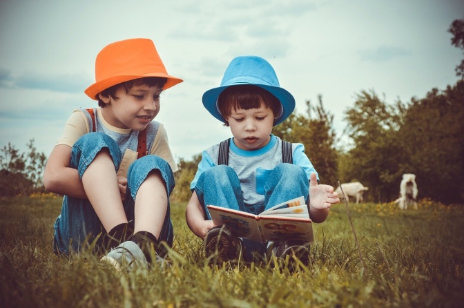 2 Jungen mit Hüten sitzen auf einer Wiese und lesen zusammen ein Buch.