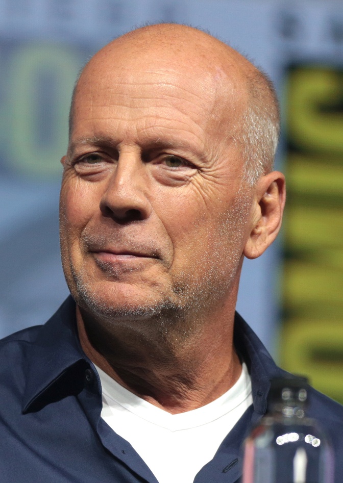 Bruce Willis mit Glatze, Dreitagebart und angedeutetem Lächeln. Er steht vor einer Logowand.