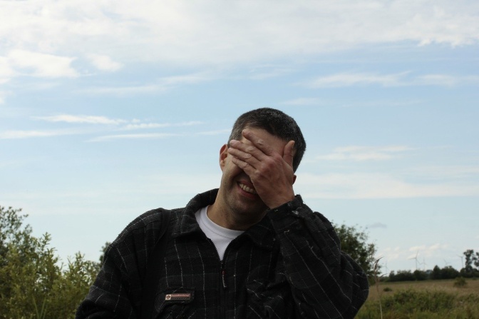 Ein Mann lacht und verdeckt sein Gesicht mit der Hand. Er steht im Freien vor einem leicht bewölkten Himmel.