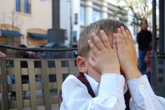 Ein blonder Junge sitzt auf einer Bank im Freien. Er versteckt sein Gesicht hinter beiden Händen.