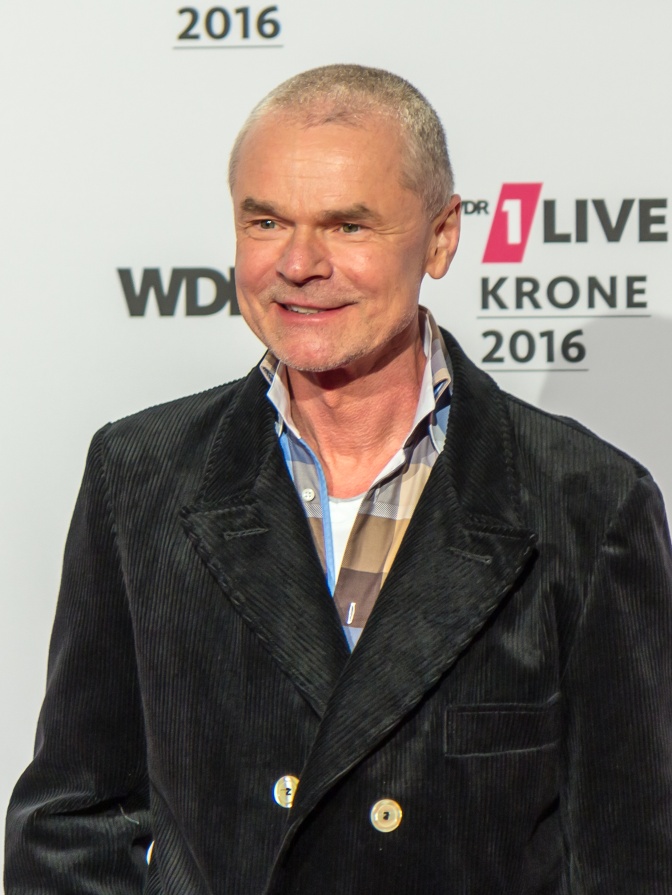 Jürgen Domian mit kurzrasierten grauen Haaren in Karohemd und Cordjacke vor einer Logowand des WDR