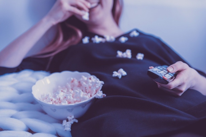 Eine Frau liegt auf der Couch und isst Popcorn. Ein Teil des Popcorns ist auf ihren Pullover gefallen. In der Hand hält sie eine Fernbedienung.