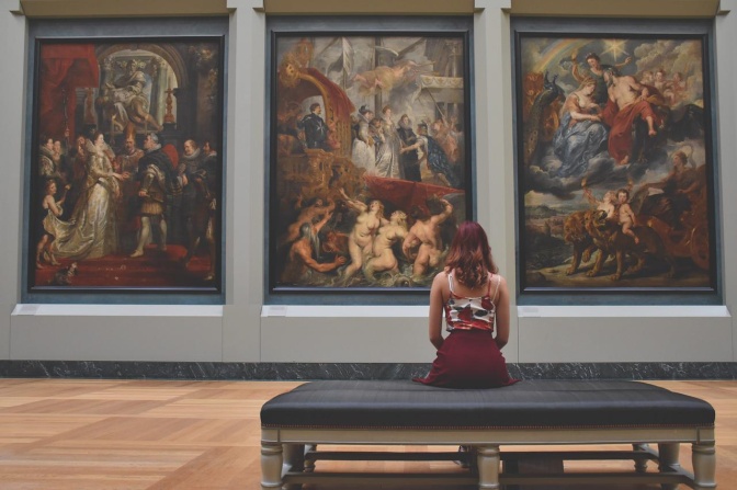 Eine Frau sitzt in einem Museum vor drei großen, monumentalen Gemälden und betrachtet sie.