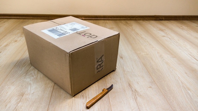 Ein Paket steht auf einem Holzboden, daneben liegt ein Cuttermesser.