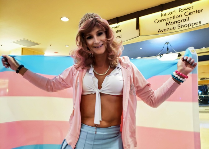 Eine trans Person hält lächelnd eine trans Flagge hinter sich ausgebreitet. Die Person trägt eine gewellte Langhaarperücke und ein Diadem.