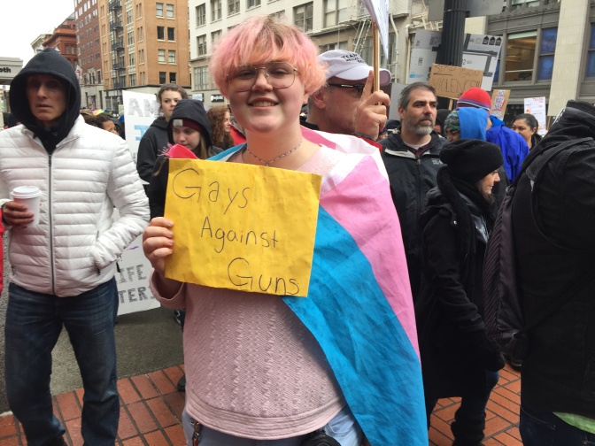 Eine trans Person mit rosanen Haaren hält ein Schild in die Kamera. Darauf steht: Gays against uns. Also übersetzt: Schwule gegen Waffen.