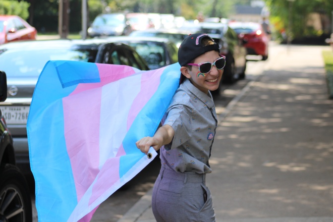 Eine männlich gelesene trans Person läuft lächelnd mit ausgebreiteten Armen und einer trans Flagge über eine Straße. Die Person trägt eine Schirmmütze und hat einen Regenbogen auf die Wange gemalt.