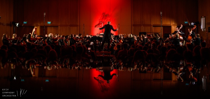 Das Orchester wird dirigiert und musiziert gemeinsam. Die Bühne ist im Hintergrund rot beleuchtet.