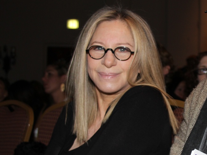 Barbra Streisand mit langen, glatten blonden Haaren in schwarzer Kleidung und mit runder, schwarzer Brille.