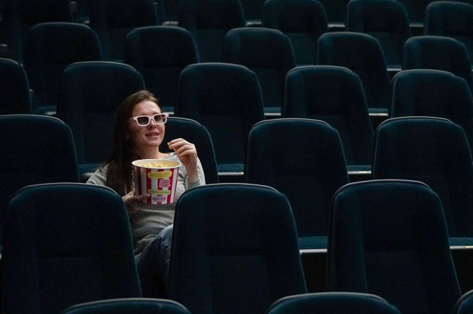 Eine Frau sitzt in einem leeren Kinosaal. Sie trägt eine 3D-Brille und isst Popcorn.