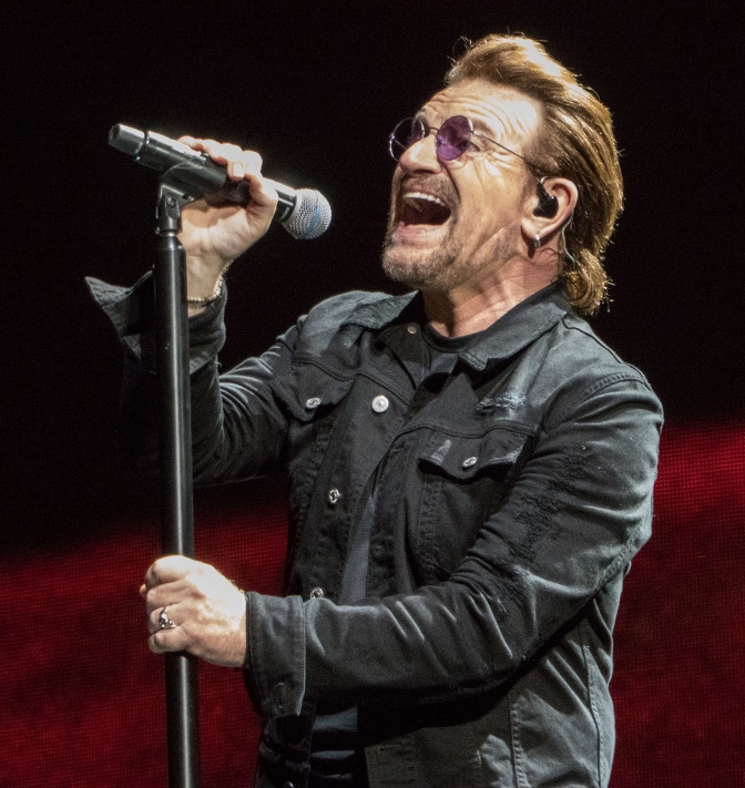 Bono singt in ein Mikro. Er steht auf der Bühne und trägt Jeansjacke und Sonnenbrille.