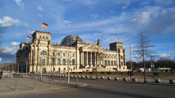 Das Bundestagsgebäude mit der Glaskuppel in der Mitte. An allen 4 Ecken des Gebäudes weht die deutsche Flagge.