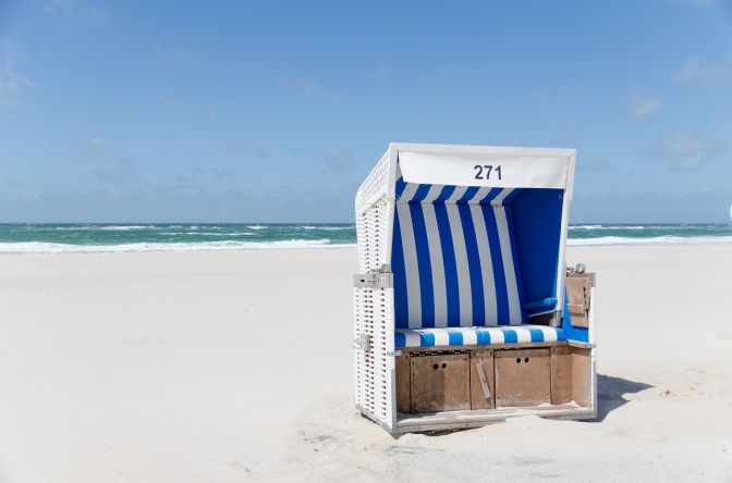 Ein einzelner weißer Strandkorb mit gestreiften Sitzkissen steht an einem ansonsten leeren Sanstrand.