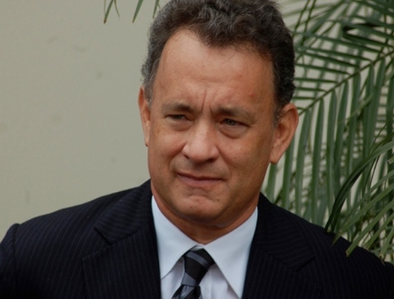 Tom Hanks mit kurzen, braunen Haaren in Anzug und Krawatte vor einer Zimmerpflanze