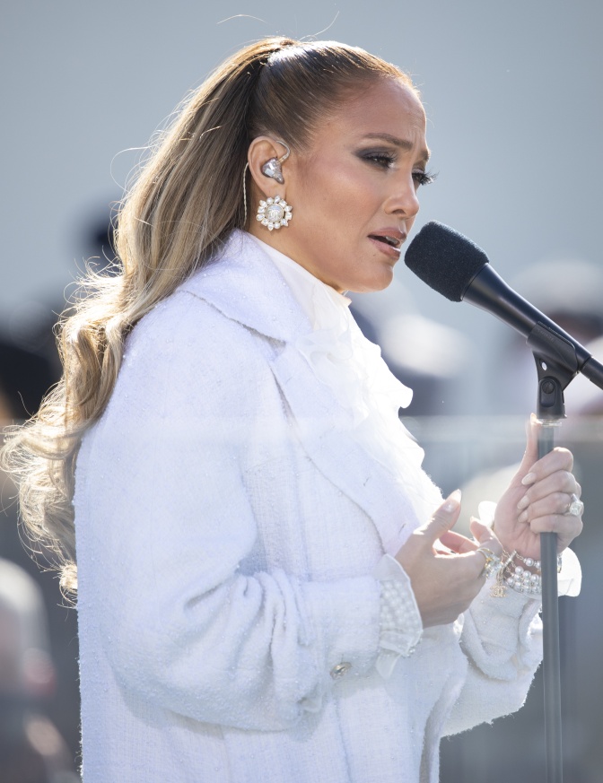 Jennifer Lopez mit langen, blonden, zum Pferdeschwanz gebundenen Haaren in einer weißen Jacke. Sie singt in ein Mikrophon.