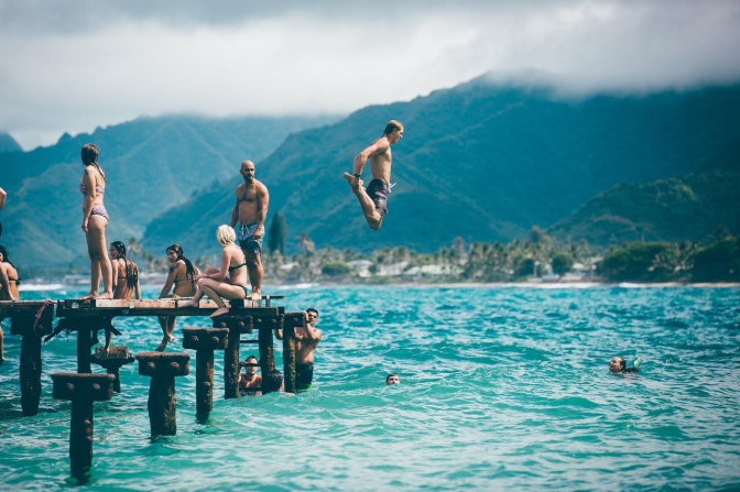 Eine Gruppe weißer Menschen in Badekleidung auf einem Holzsteg an einem Strand. Ein Mann springt gerade vom Steg aus ins Wasser.