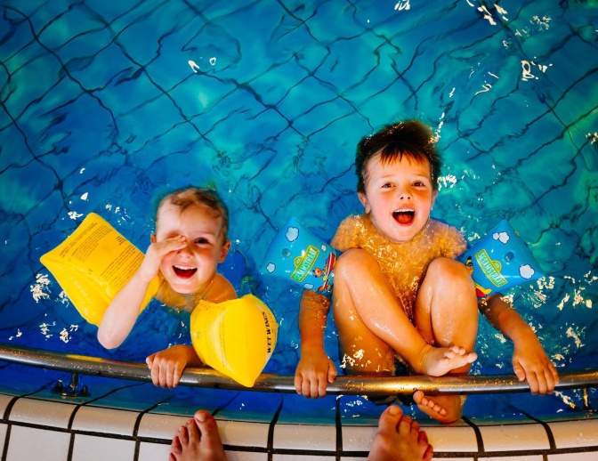 2 Kinder mit Schwimmflügeln am Beckenewand eines Schwimmbads