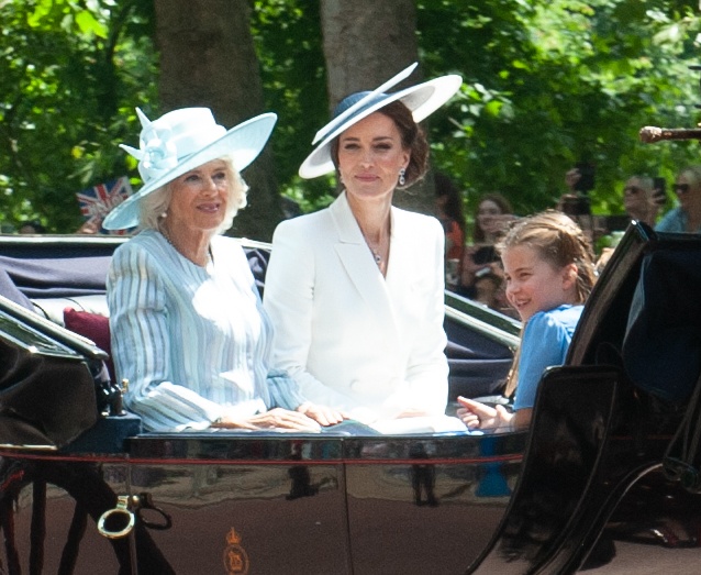 Camilla und Kate sitzen zusammen in einer offenen Kutsche. Beide lächeln und tragen große Hüte mit breiten Krempen. Auf der anderen Seite der Kutsche sitzt Kates Tochter Charlotte.