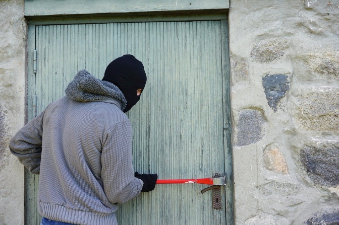 Ein Einbrecher mit schwarzer Strumpfmaske bricht mit einem Stemmeisen eine Tür auf.