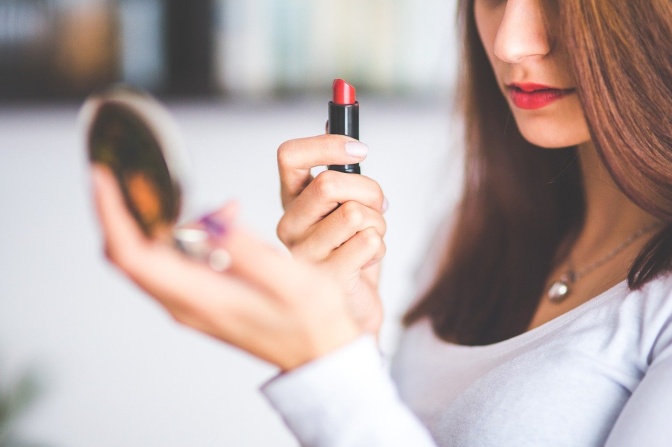 Eine Frau hält einen Schminkspiegel in der einen, einen roten Lippenstift in der anderen Hand.