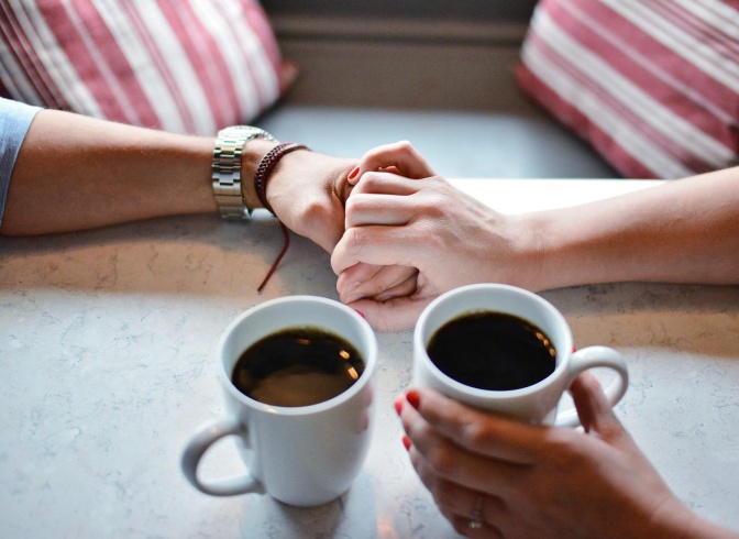 Zwei Personen halten Händchen. Ihre ineinander verschlungenen Hände liegen neben zwei Tassen Kaffee auf dem Tisch.