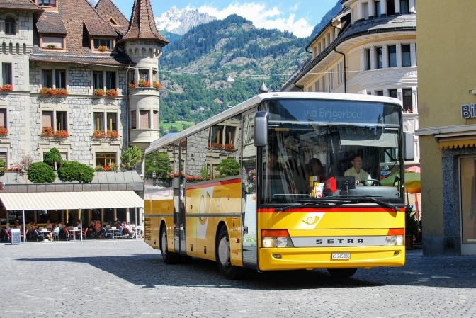 Ein gelber Bus mit dem Logo der Post fährt durch eine malerische Altstadtkulisse.