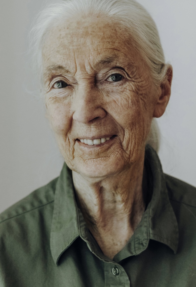 Jane Goodall mit weißen, zurückgebundenen Haaren in einer khakifarbenen Bluse. Sie lächelt in die Kamera.