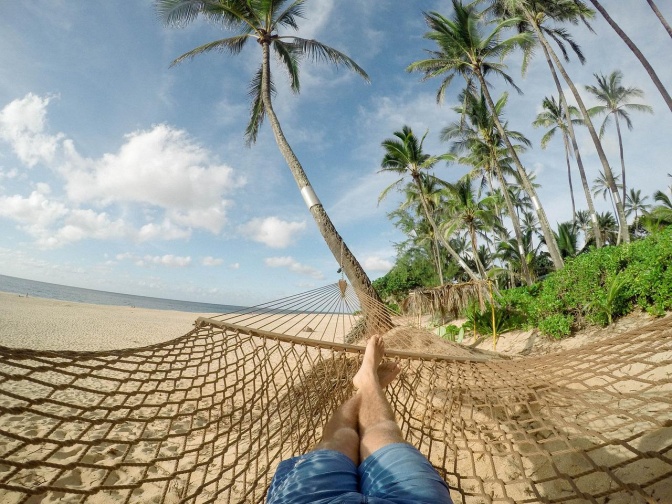 Eine Person liegt in einer Hängematte, man sieht nur ihre Beine und nackten Füße. Im Hintergrund sieht man Strand und Palmen.