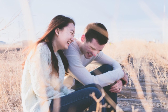 Ein Mann und eine Frau sitzen eng nebeneinander in einem Kornfeld und lachen. Er sieht europäisch aus, sie asiatisch.