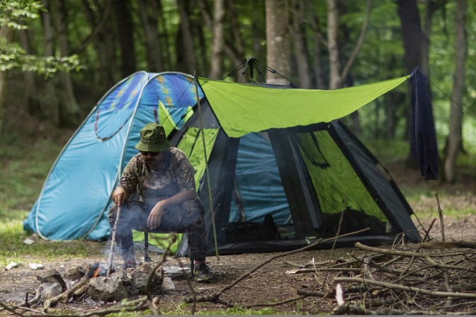 Ein Mann sitzt vor einem Zelt im Wald und stochert mit einem stock in einem Lagerfeuer herum.