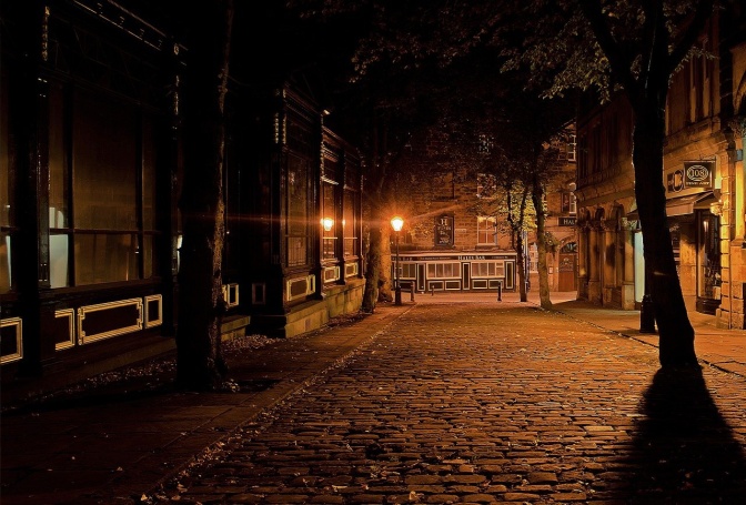 Eine Straße mit Kopfsteinpflaster bei Nacht. Am Ende der Straße leuchtet eine einzelne Straßenlaterne. Rechts und links stehen Bäume.