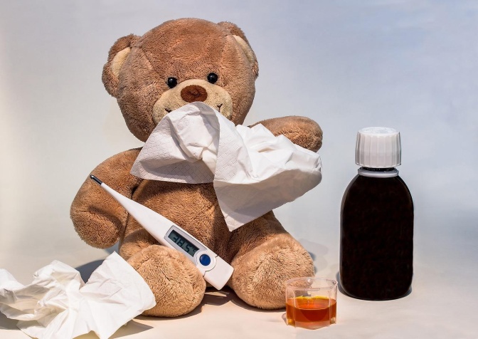 Ein Teddybär mit Fieberthermometer, Hustensaft und einem Taschentuch.